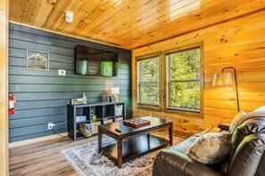 A cozy living room with a dark green wall, a wooden ceiling, a leather sofa, a coffee table, and a fireplace. - NEW! Modern Smokies Cabin! Hot Tub! Games! POOL! (Pigeon Forge)