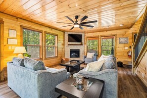 L’intérieur d’un chalet accueillant, avec une cheminée, deux canapés, une table basse et un ventilateur de plafond.