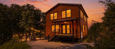 A two-story wooden cabin with a porch, surrounded by trees and string lights.