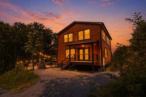 Une cabane en bois à deux étages, dotée d’un porche et entourée d’arbres et de guirlandes lumineuses.