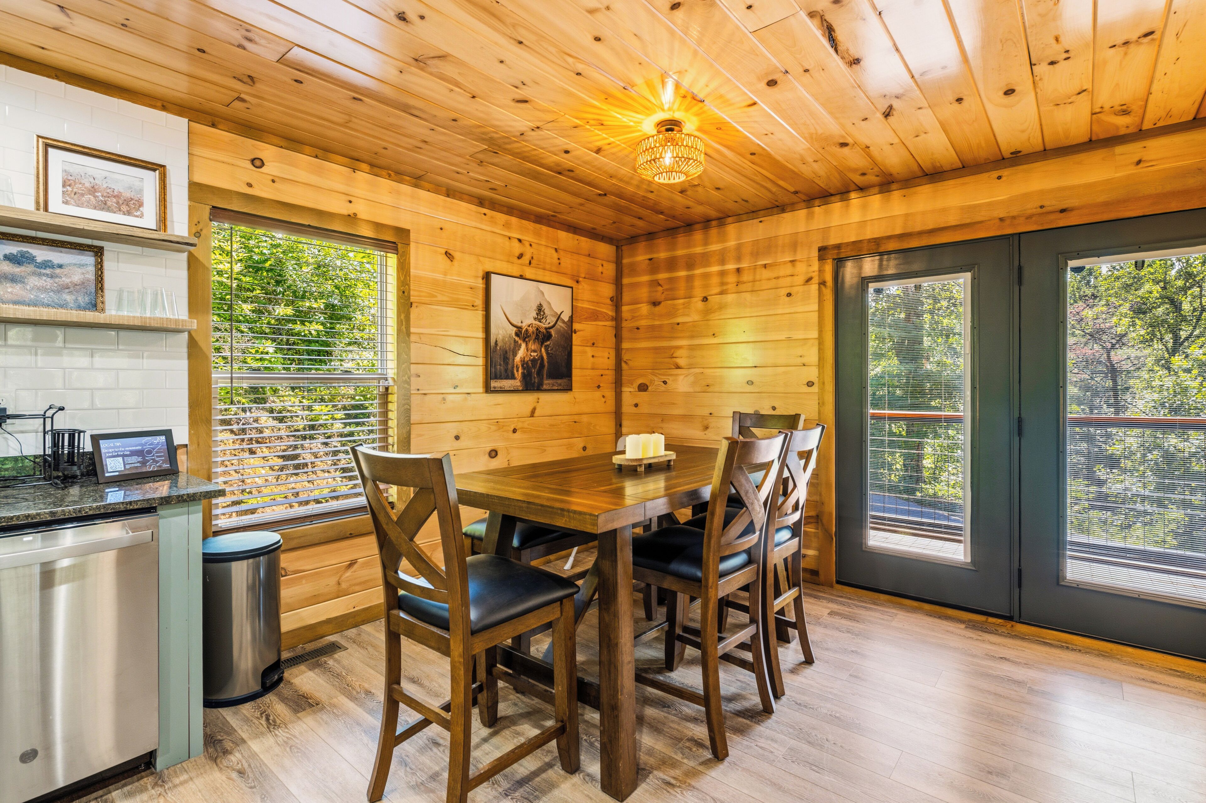 Interior de madera con una mesa de comedor, sillas, una zona de cocina y un amplio ventanal con persianas.