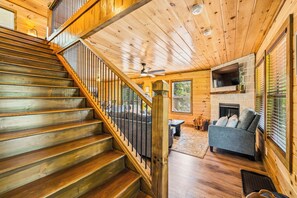 A wooden staircase leading to a living area with a fireplace, a sofa, and a television mounted on a stone wall. - NEW! Modern Smokies Cabin! Hot Tub! Games! POOL! (Pigeon Forge)