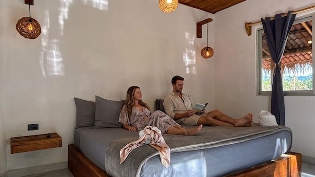 Ein Schlafzimmer mit Holzboden, einem Bett mit grauer Bettwäsche und zwei Personen, die darauf sitzen.
