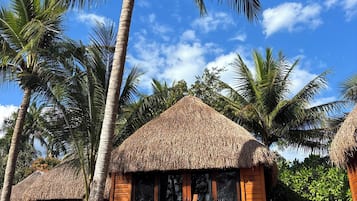 A wooden bungalow with a thatched roof, surrounded by palm trees, and a clear blue sky.