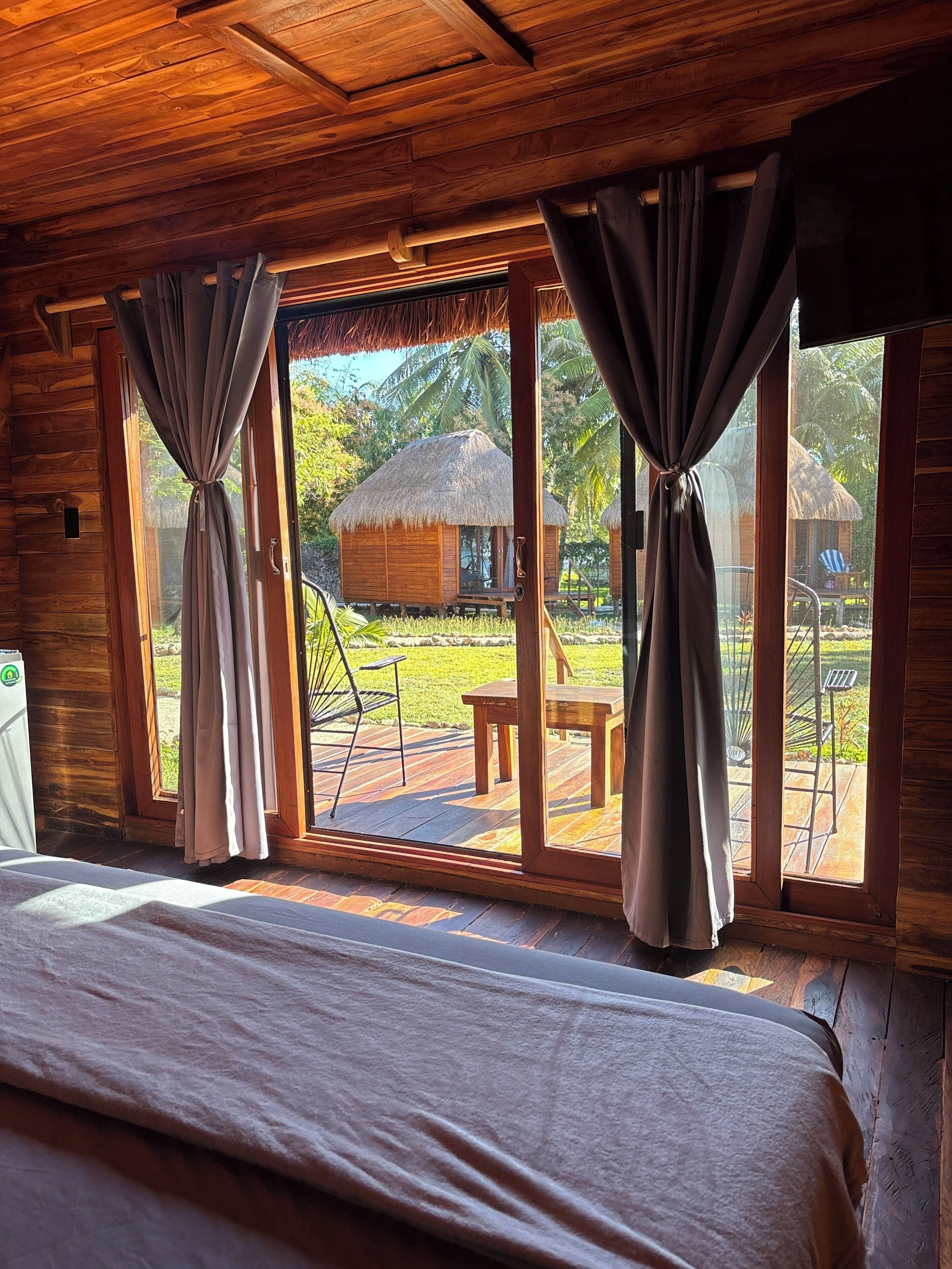 A wooden room with a bed, curtains, and large windows overlooking a thatched-roof structure and a grassy area.