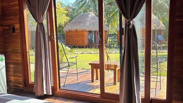 A wooden room with a bed, curtains, and large windows overlooking a thatched-roof structure and a grassy area.