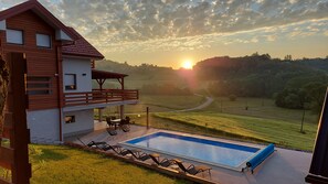 A house with a pool, outdoor furniture, and a view of the sunset over a hilly landscape.