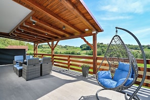 A covered outdoor seating area with wicker furniture and a hanging chair, overlooking a scenic landscape.