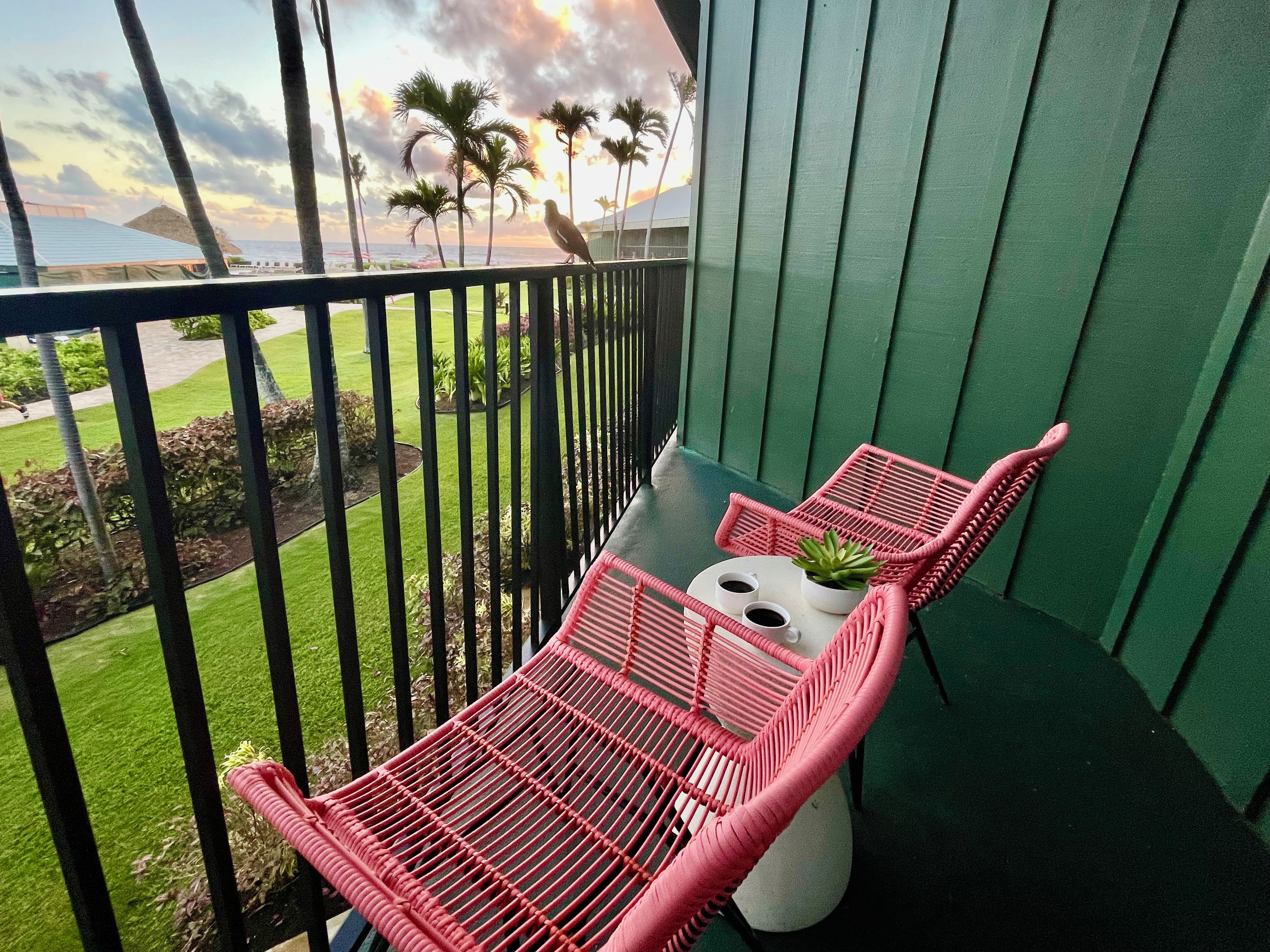 A balcony with a green railing, a pink chair, a small table with two cups of coffee, and a potted plant.