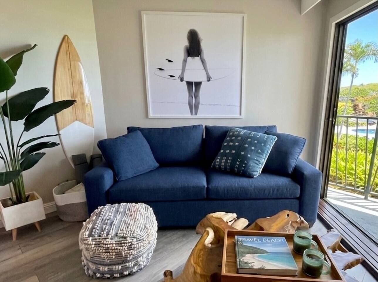 A living room with a blue sofa, a framed artwork, a surfboard, and a view of the outdoors.