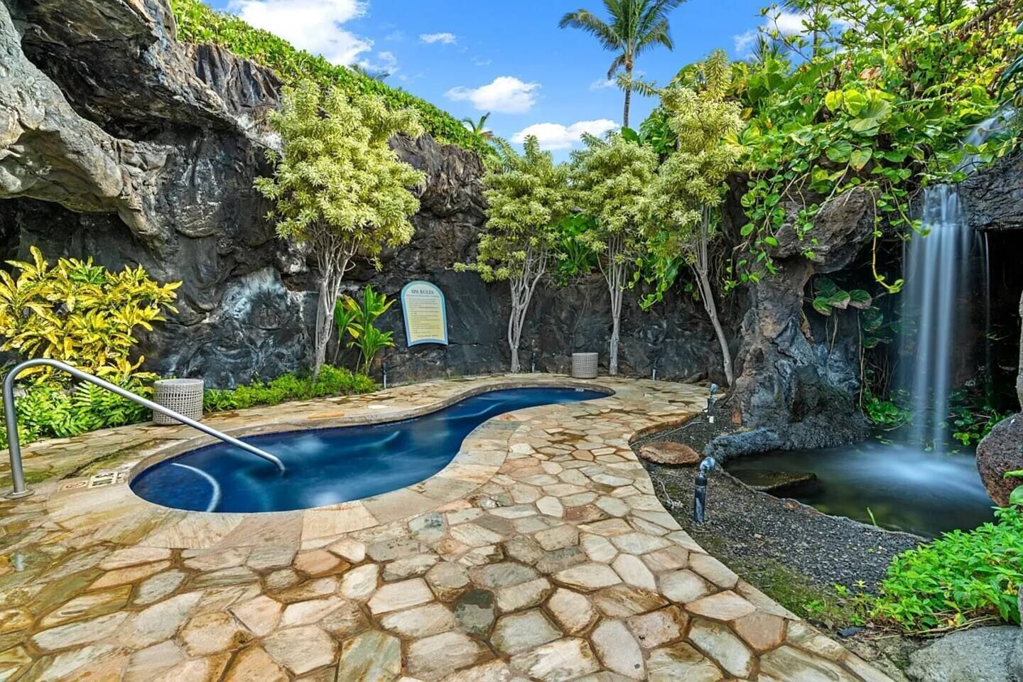 A spa pool with a stone pathway, surrounded by tropical vegetation and a waterfall.