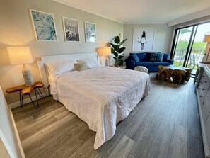 A bedroom with a large bed, a nightstand, a lamp, a blue sofa, and a view of the outdoors.