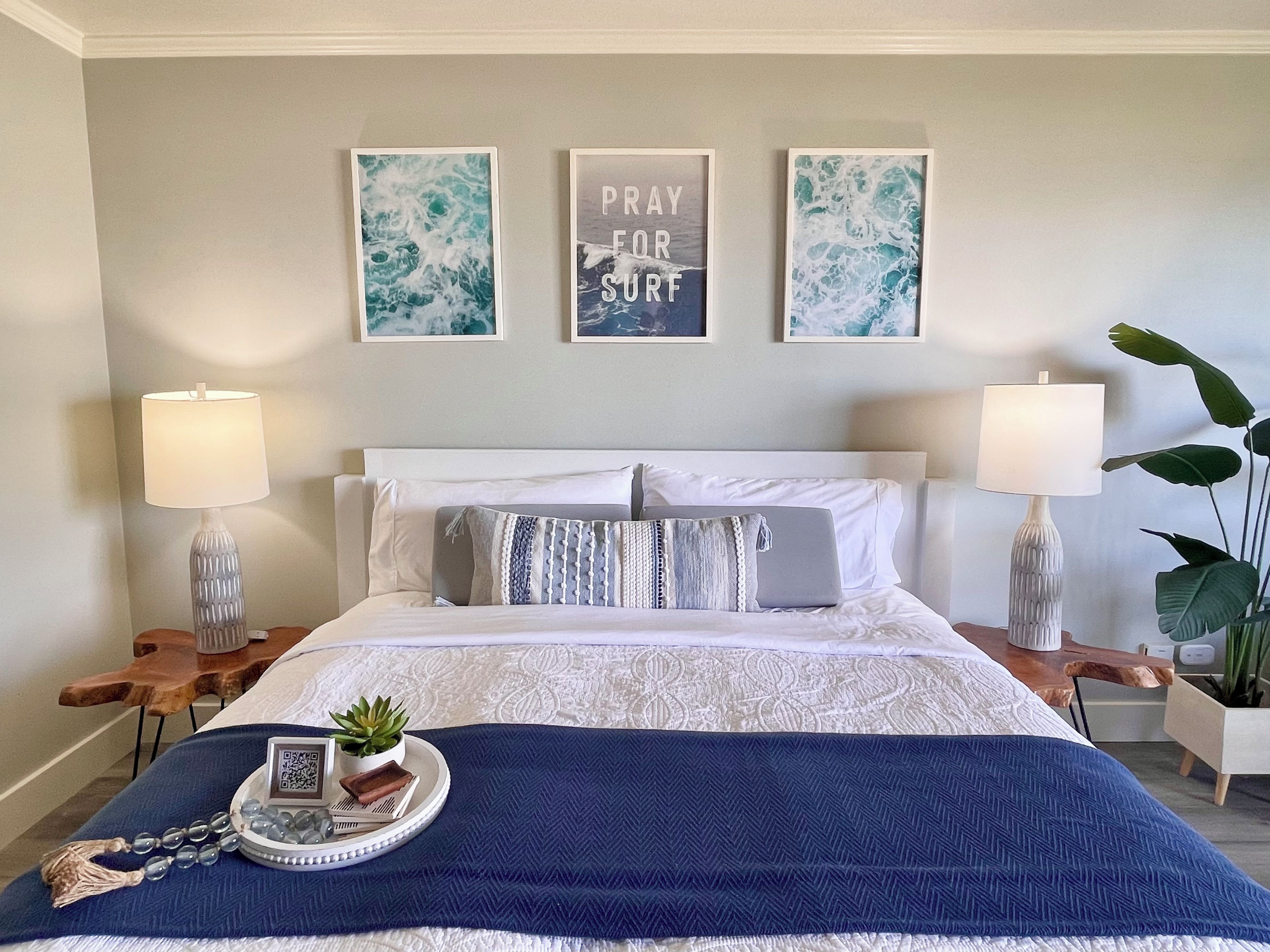 Une chambre à coucher bien rangée, avec un lit, deux tables de chevet, un plaid bleu, une petite plante et trois tableaux encadrés accrochés au mur.