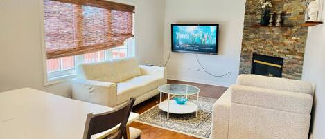 A modern living room with a white sofa, a glass coffee table, a dining table with chairs, a TV mounted on the wall, and a stone fireplace.