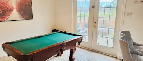 Ein Billardtisch in einem Raum mit einer zur Außenseite führenden Tür, einem Sessel und einem Gemälde an der Wand.