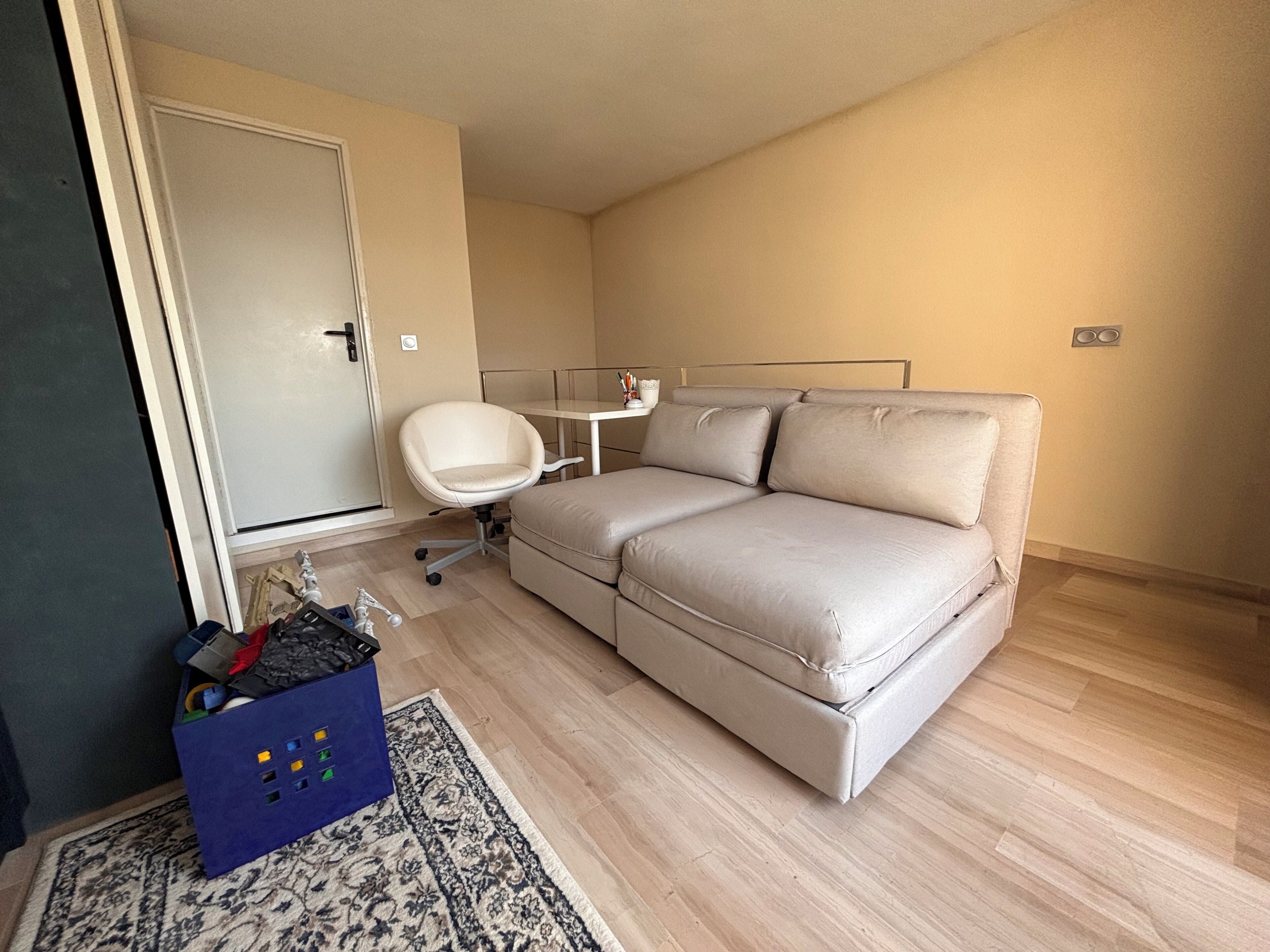 Una habitación pequeña amoblada con sofá, una silla, escritorio y puerta.