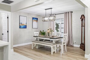 A dining area with a wooden table, white chairs, a bench, a chandelier, and a large window with curtains.