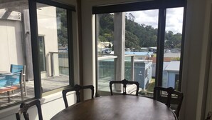 A dining area with a round table and four chairs, large windows with a view of buildings and trees, and a balcony with a chair and table. - Absolute beachfront in Waiwera (Waiwera)