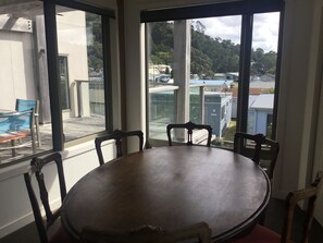 A dining area with a round table and four chairs, large windows with a view of buildings and trees, and a balcony with a chair and table. - Absolute beachfront in Waiwera (Waiwera)