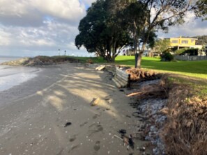 A sandy beach with footprints, a grassy area, and a yellow building in the background. - Absolute beachfront in Waiwera (Waiwera)
