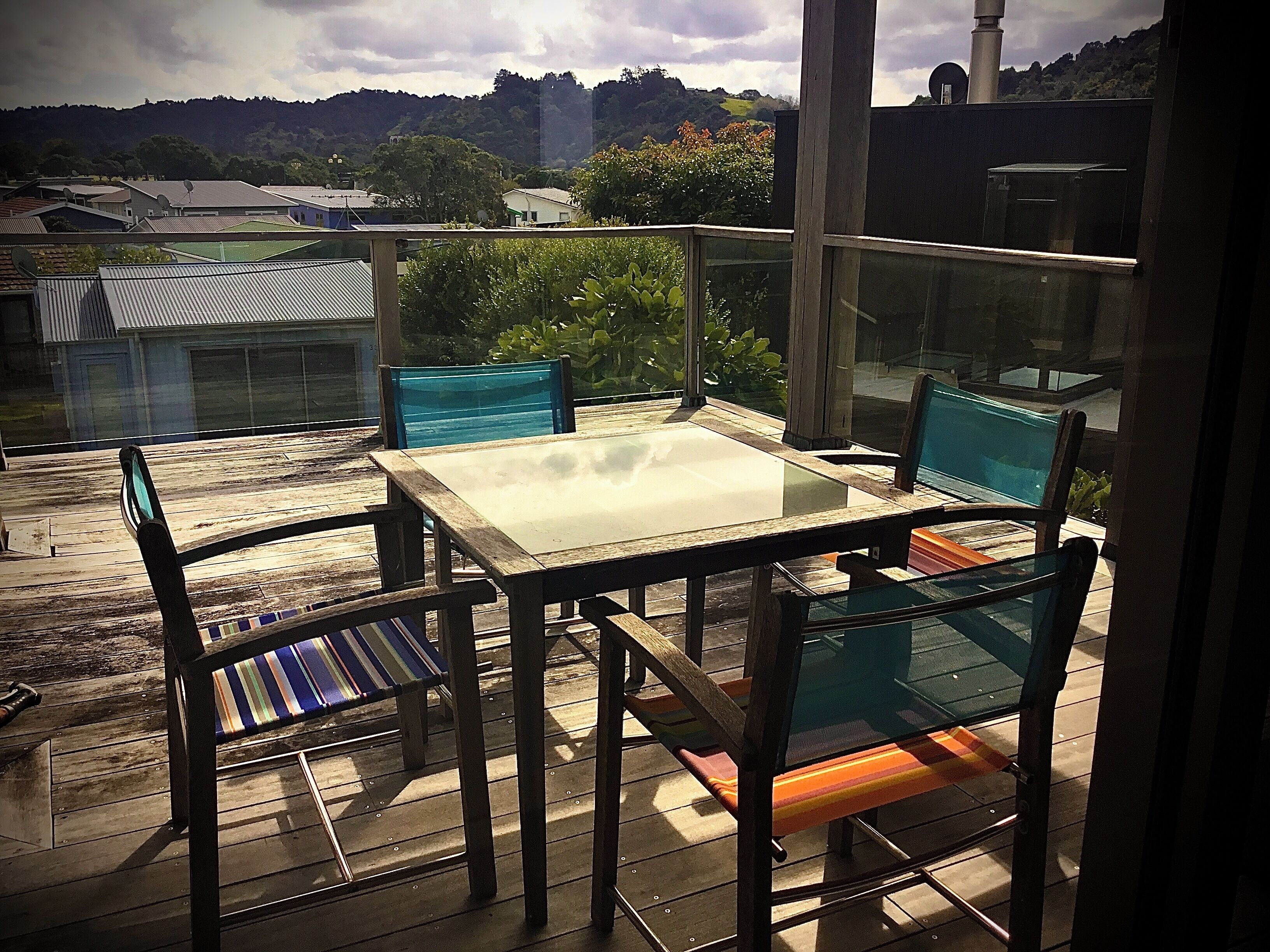 A balcony with a glass table and chairs, overlooking a residential area with houses and greenery.