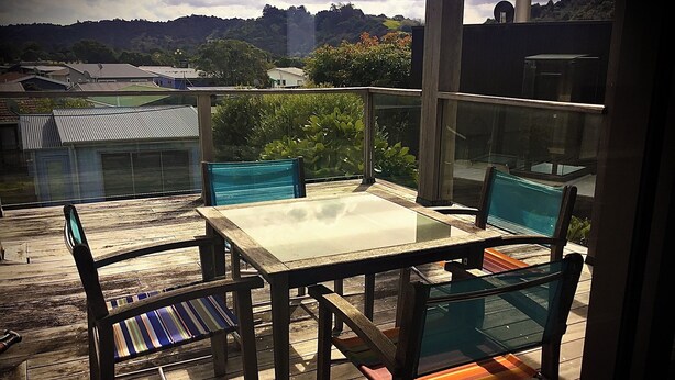 A balcony with a glass table and chairs, overlooking a residential area with houses and greenery. - Absolute beachfront in Waiwera (Waiwera)