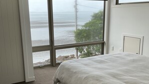 A bedroom with a large bed, a wooden nightstand, a window with a view of the sea, and a ceiling light. - Absolute beachfront in Waiwera (Waiwera)
