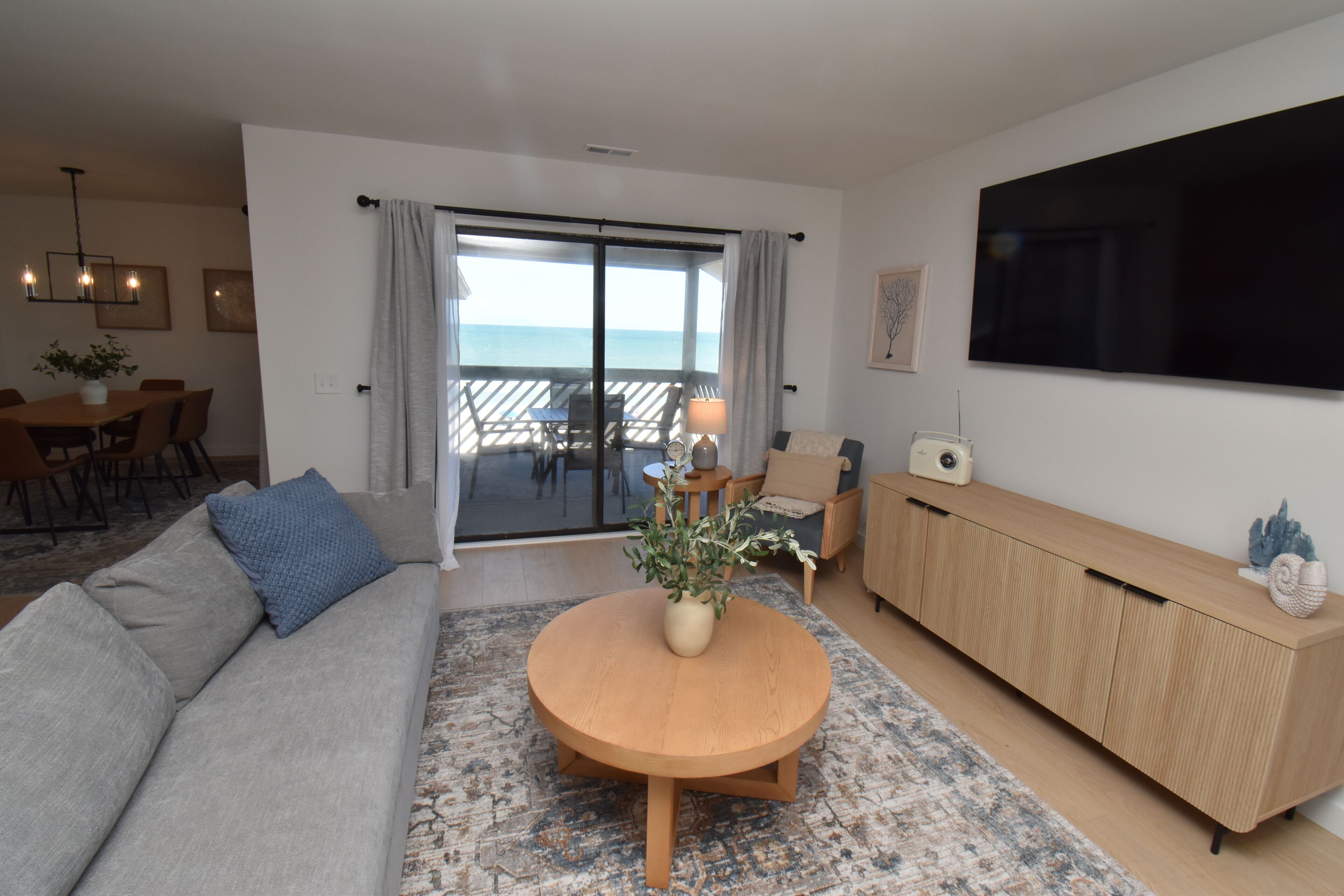 Uma sala de estar moderna com um sofá, uma mesa de centro redonda de madeira, uma televisão e uma varanda com vista para o mar.