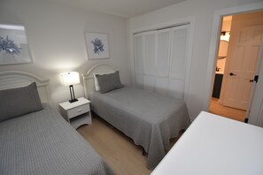 Une chambre avec deux lits, une table de chevet avec une lampe, une armoire et une porte donnant sur une autre pièce.