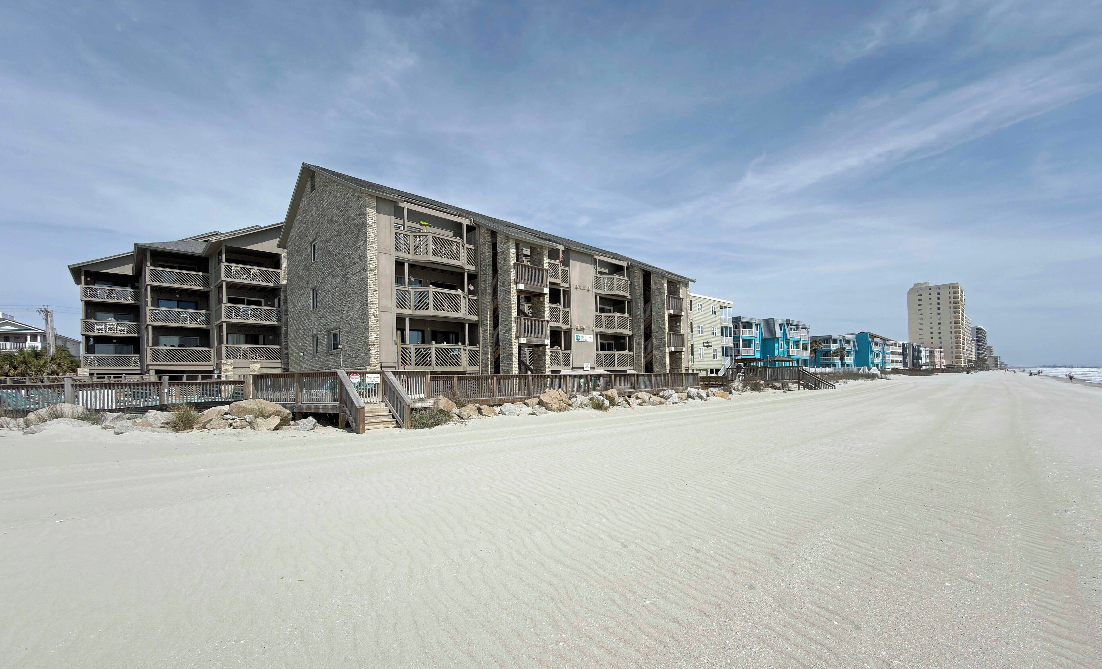 Un hotel frente a la playa con varios balcones, una playa arenosa en primer plano y un cielo despejado.