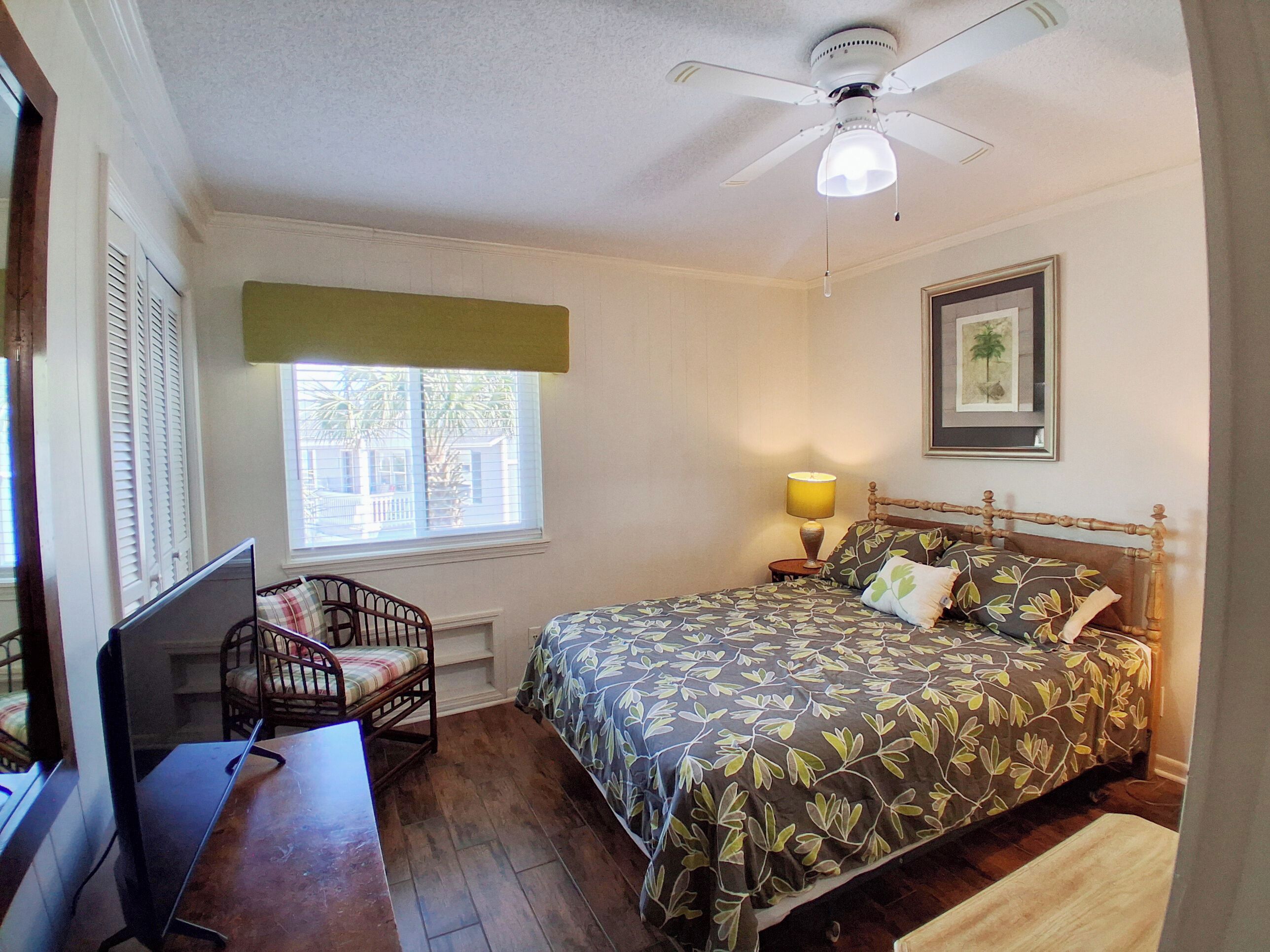 A bedroom with a bed, a desk, a chair, a window with shutters, a ceiling fan, and a wall-mounted lamp.
