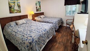 A bedroom with two beds, a ceiling fan, paintings on the wall, a window with blinds, and a television on a stand. - South Pointe Villas 6 - Oceanfront Complex (Oceanview Unit) in Garden City Beach (Garden City)