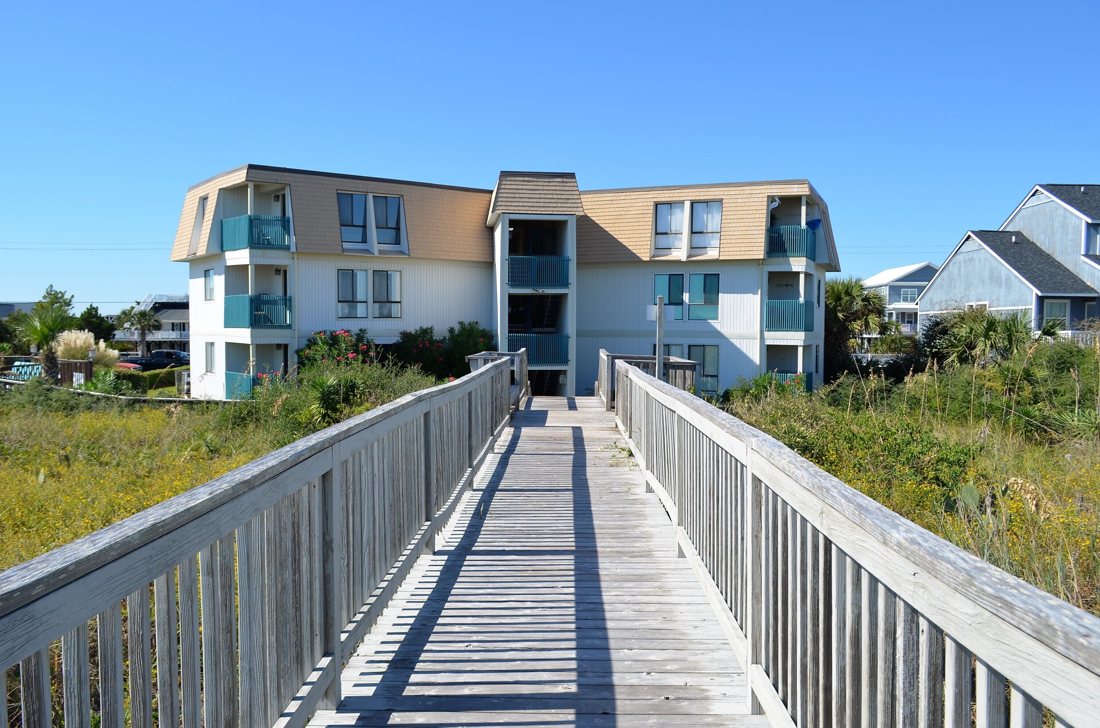 A wooden boardwalk leading to a multi-story building with balconies and a clear sky.