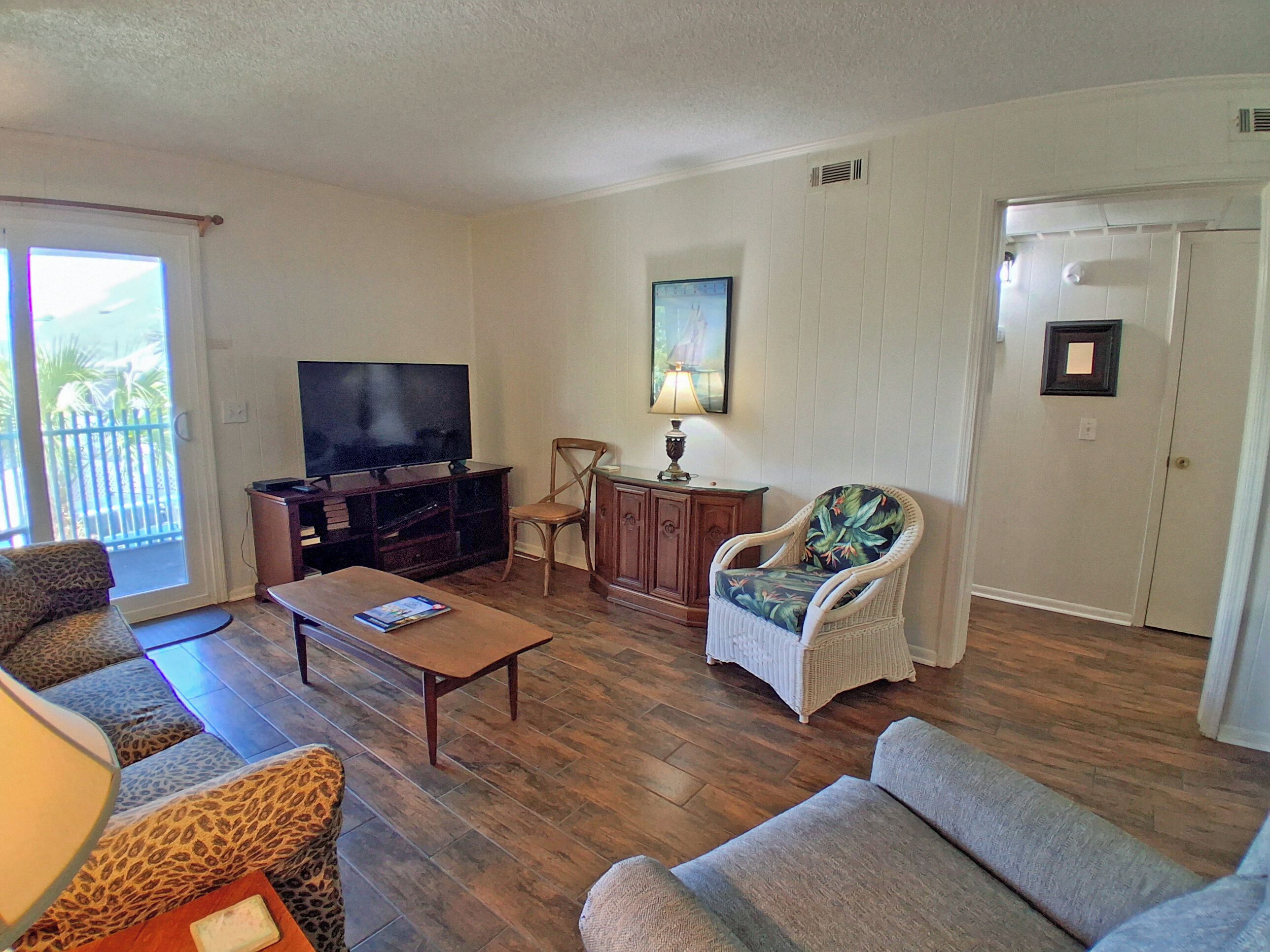 A living room with a flat-screen TV, a wooden coffee table, two sofas, a wicker armchair, and a door with a small picture frame.