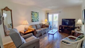 A living room with a sofa, armchair, coffee table, television, and a balcony door. - South Pointe Villas 6 - Oceanfront Complex (Oceanview Unit) in Garden City Beach (Garden City)