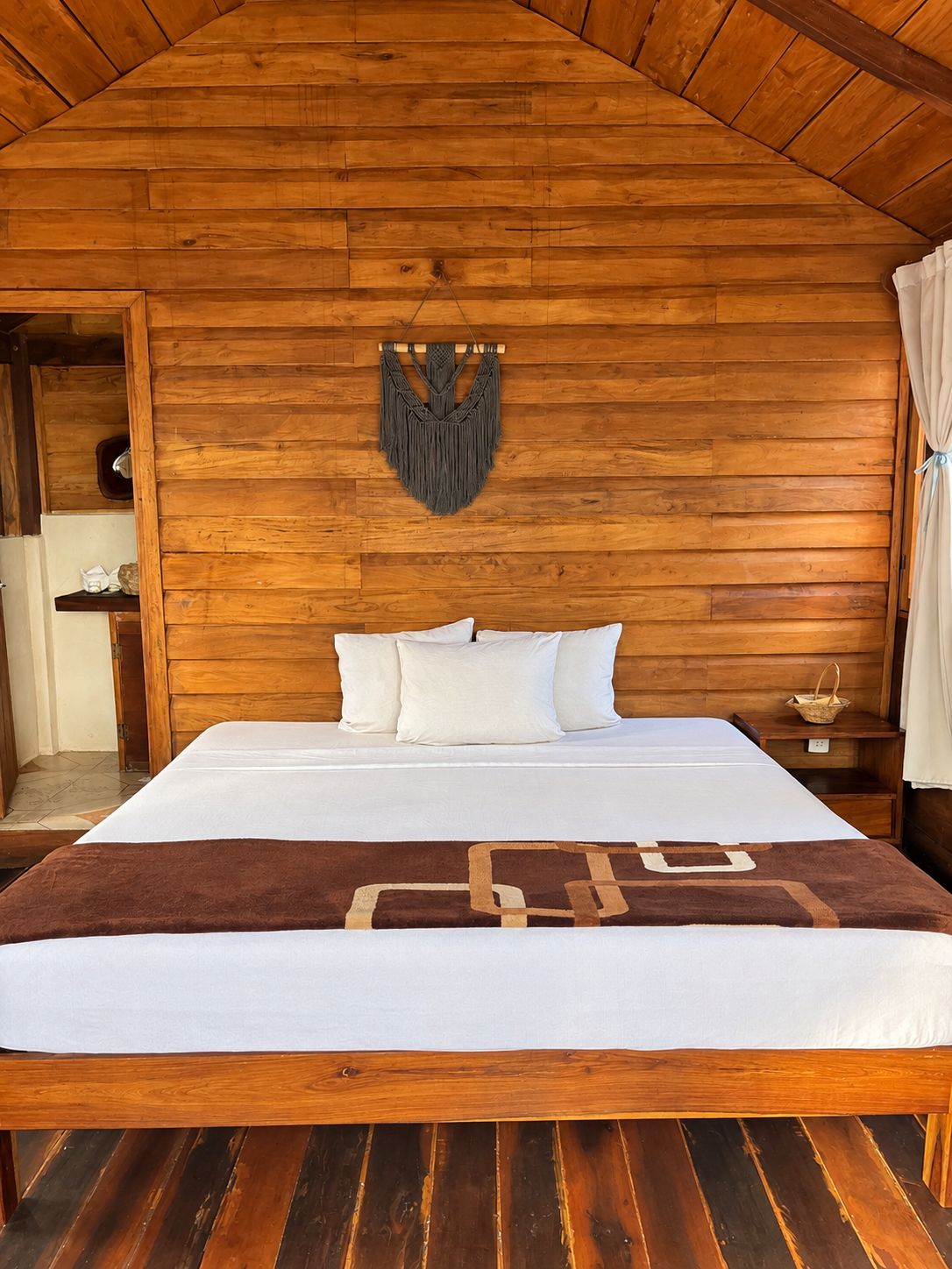 A wooden-framed room with a bed, a macrame wall hanging, and a wooden nightstand.