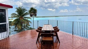 Mesa y sillas de madera en un balcón con vistas a un mar azul cristalino y palmeras.