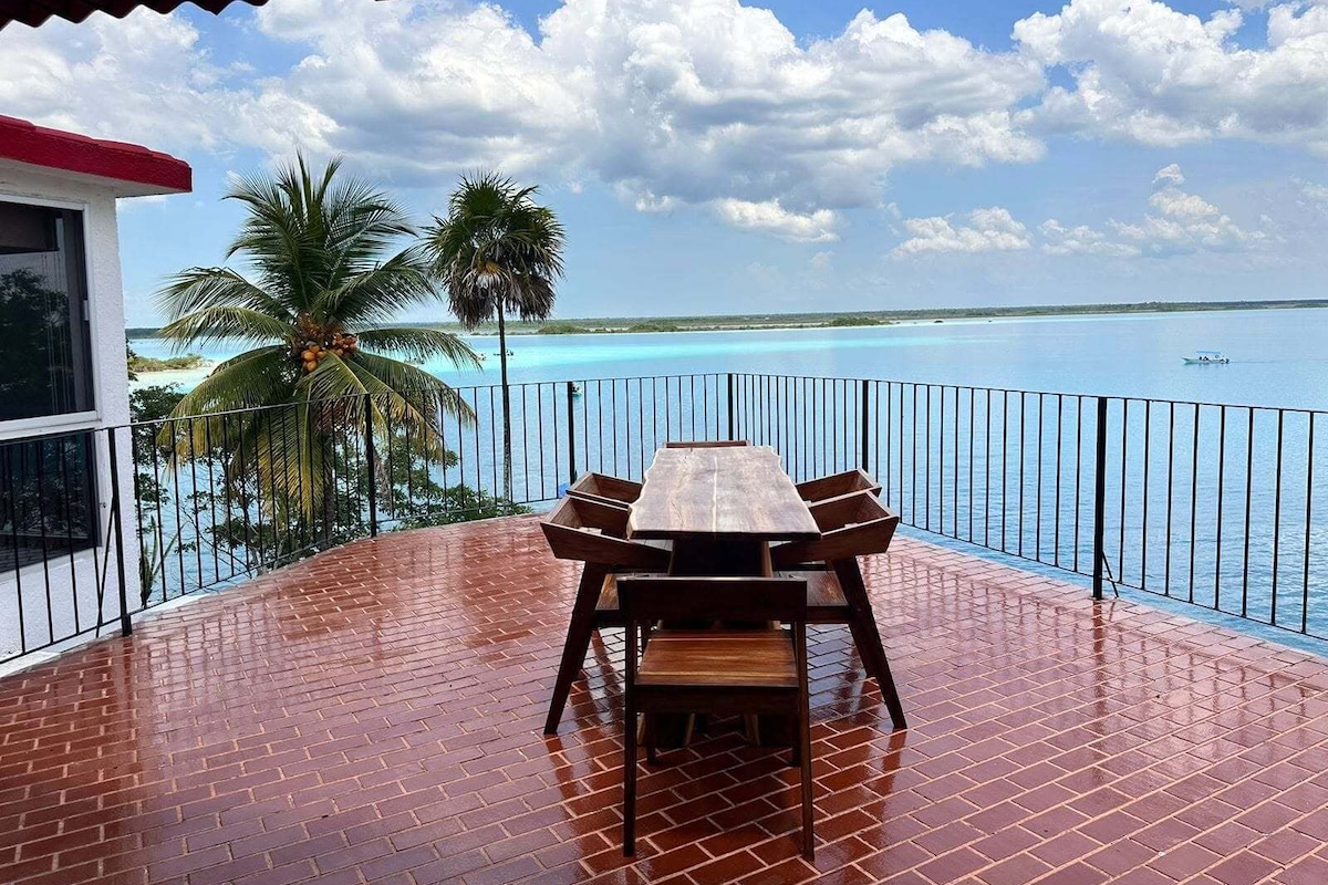 Mesa y sillas de madera en un balcón con vistas a un mar azul cristalino y palmeras.