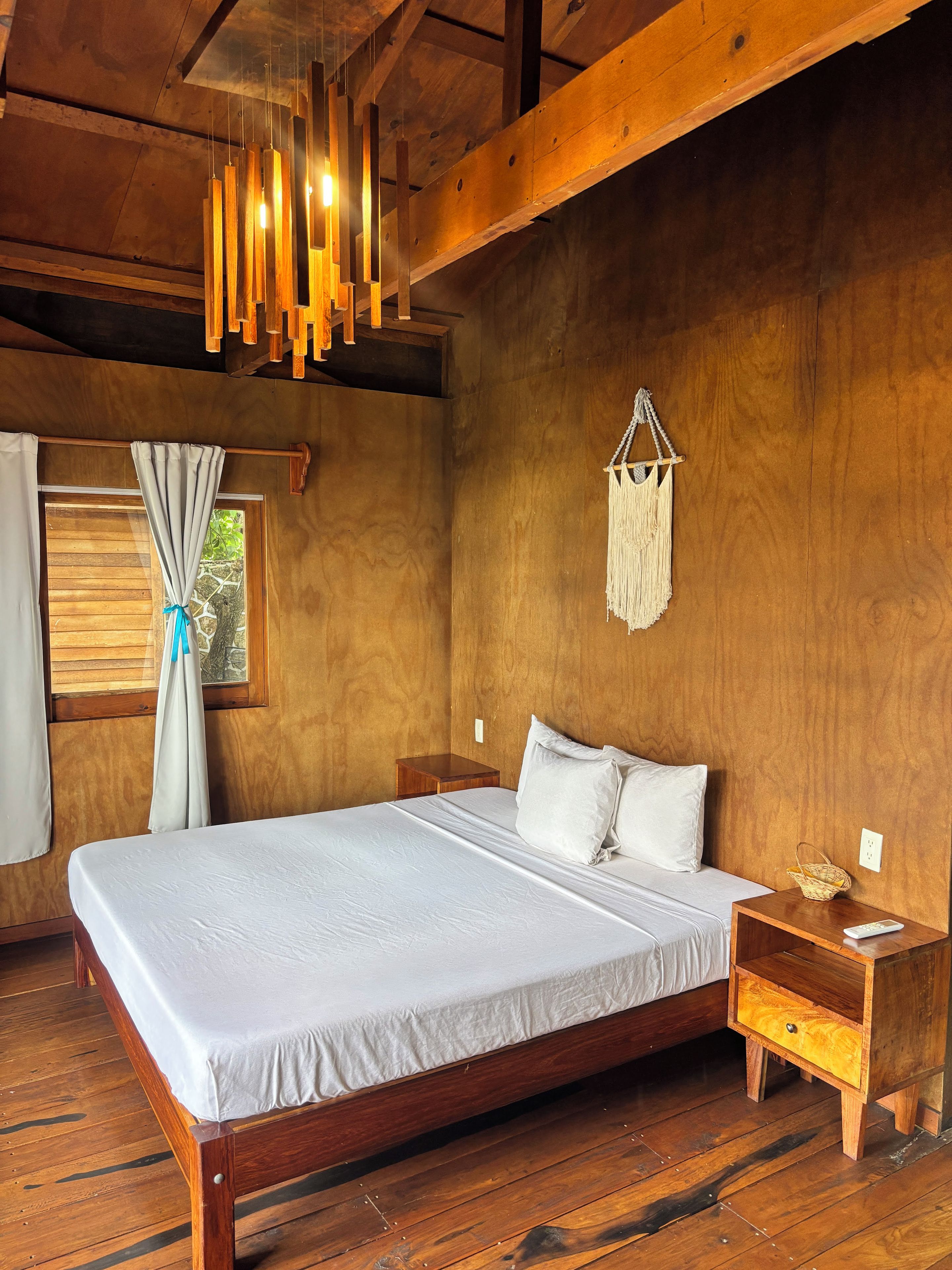A wooden room with a bed, nightstands, a hanging light fixture, and a window with curtains.