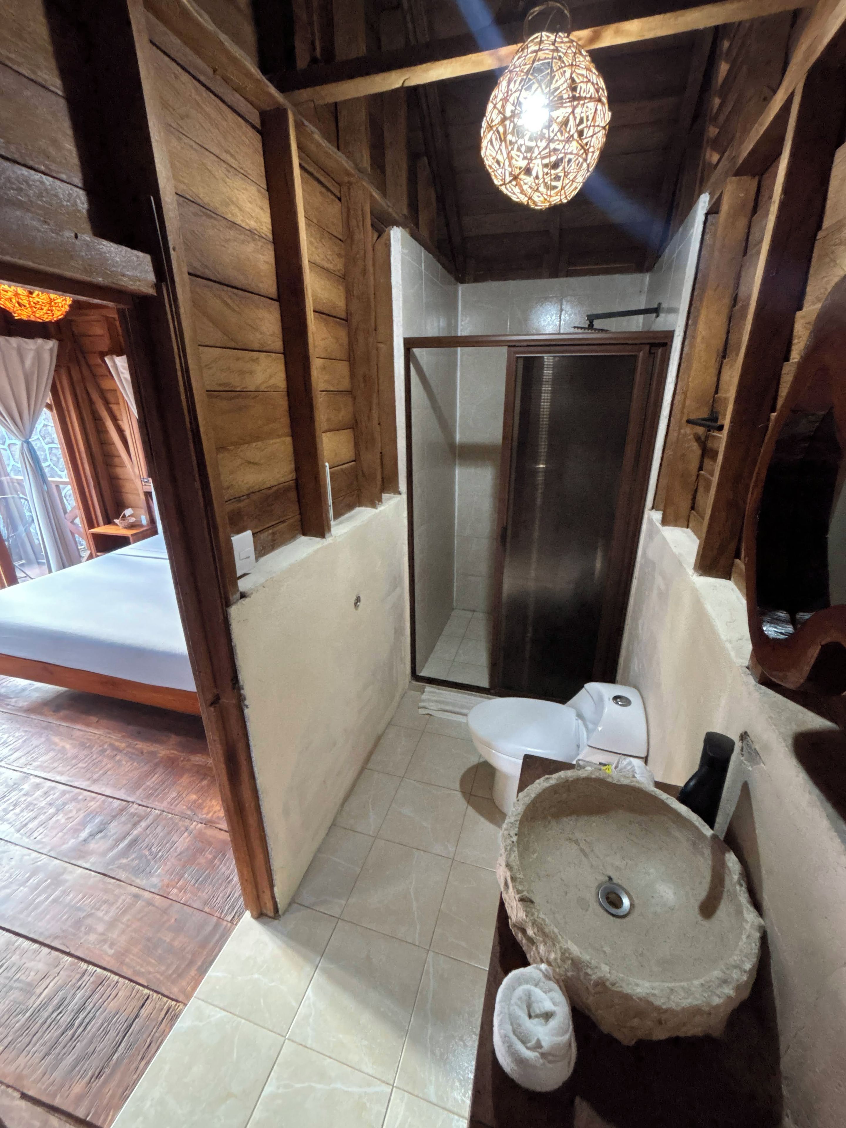 A compact bathroom with a shower, a sink with a stone basin, and a bed visible through a doorway.