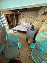 Un dormitorio con pared de piedra, una cama con ropa de cama blanca, dos sillas turquesas y una mesita de noche con lámpara.
