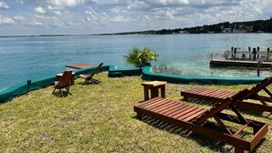 Un espace verdoyant avec des chaises longues en bois et une petite table, offrant une vue sur une étendue d’eau calme, avec un quai en arrière-plan.