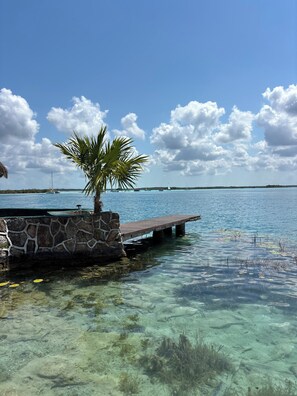 Ein klarer, blauer Himmel, eine Palme, eine Steinmauer und ein hölzerner Steg, der sich in ein ruhiges Meer erstreckt.