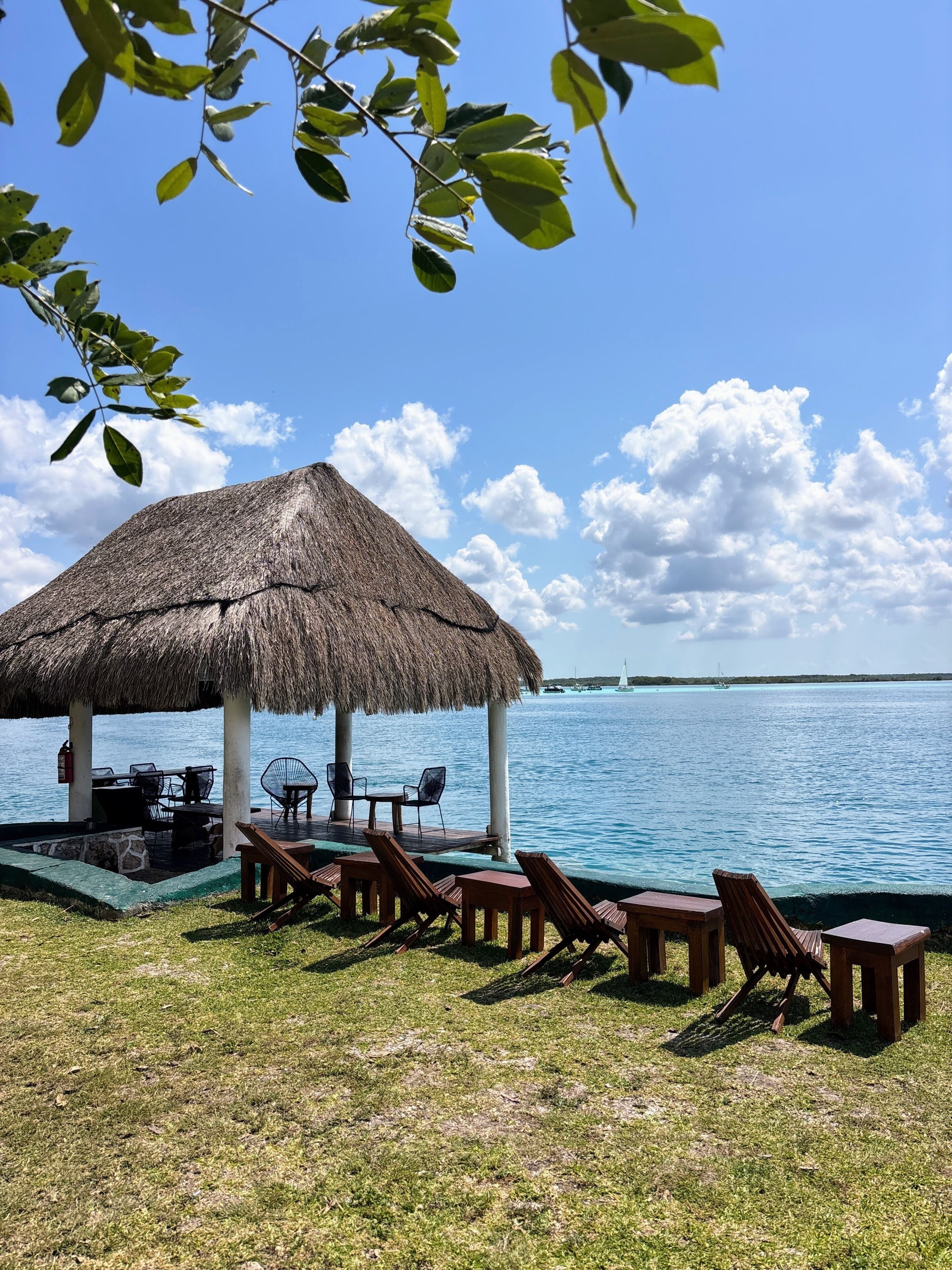 A thatched-roof outdoor seating area with lounge chairs and a table, overlooking a calm body of water.