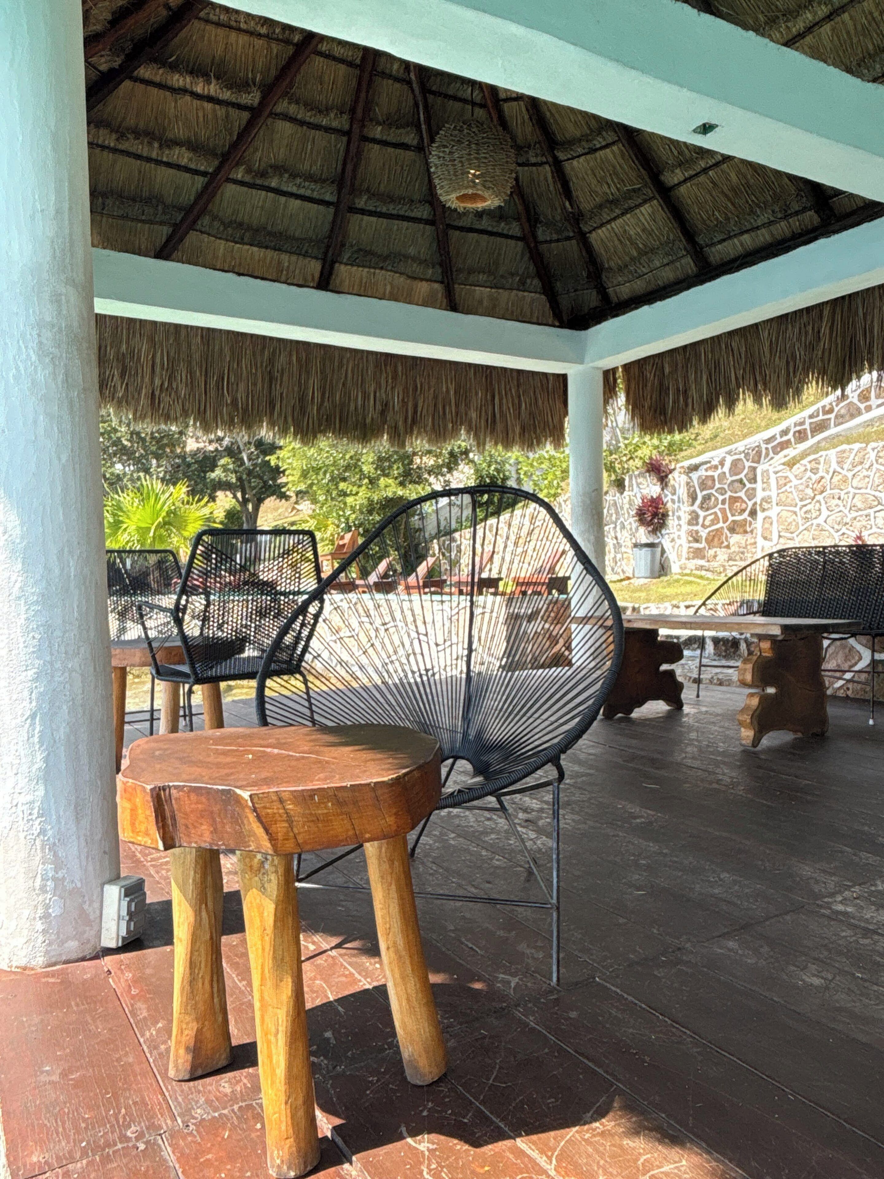Un patio exterior con techo de paja, mobiliario de madera y pared de piedra.