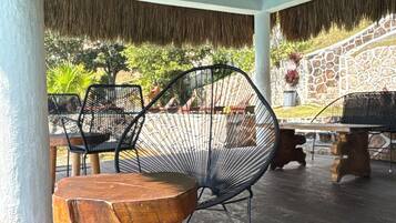 Un patio exterior con techo de paja, mobiliario de madera y pared de piedra.