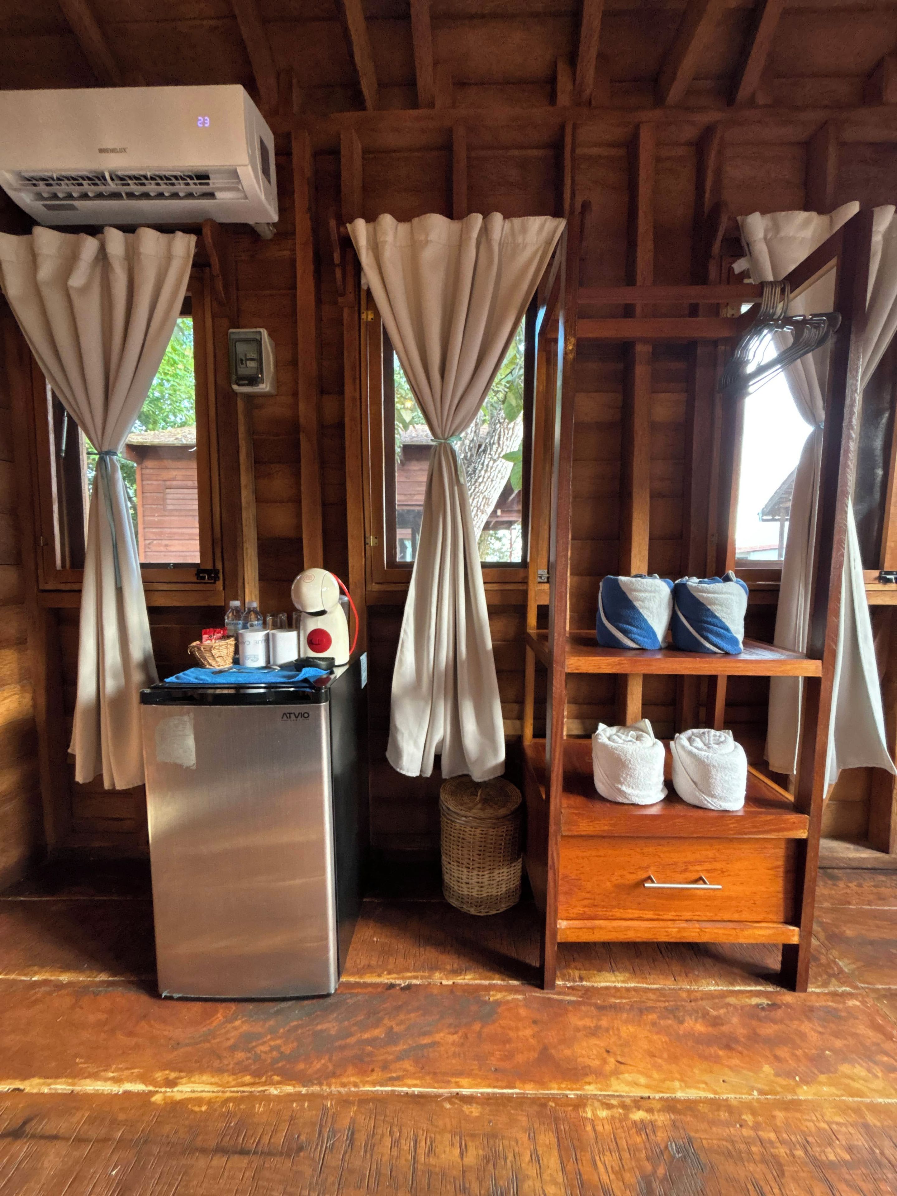 Interior de una cabaña de madera con kitchenette, un refrigerador pequeño, un estante con toallas y cortinas atadas en la parte superior.