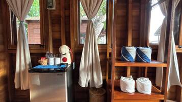 Interior de una cabaña de madera con kitchenette, un refrigerador pequeño, un estante con toallas y cortinas atadas en la parte superior.