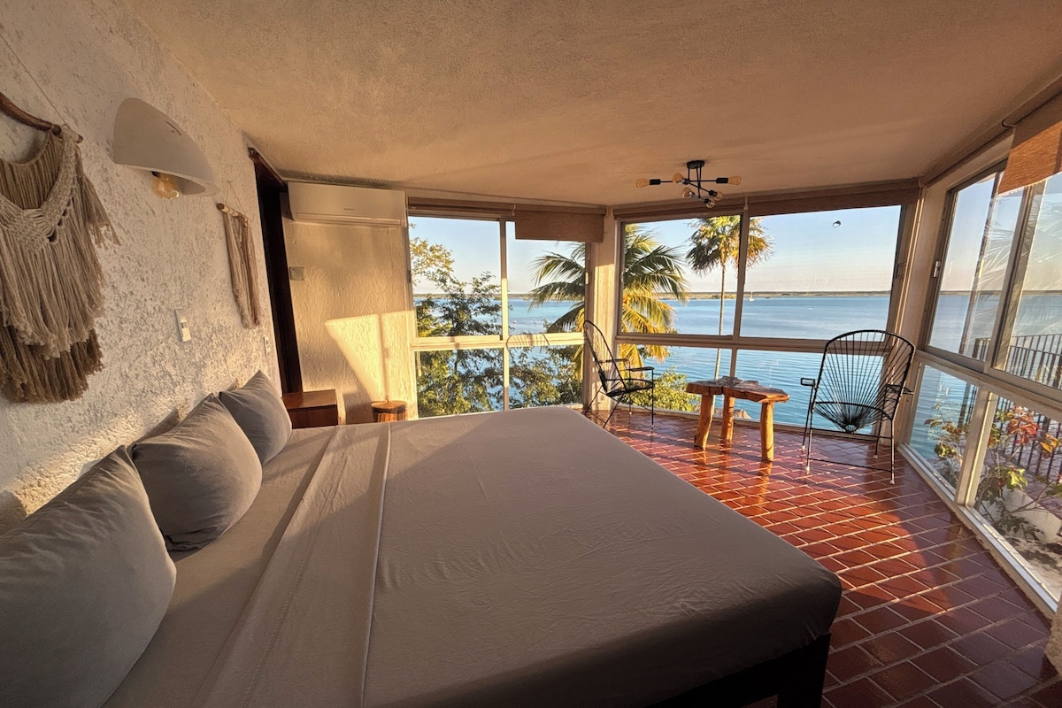 Un dormitorio con una cama grande, una mesita de noche, una lámpara y vistas al mar a través de amplias ventanas.