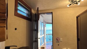 Une salle de bain avec un lavabo en pierre, des toilettes, un panier en osier et une vue sur la mer par une porte ouverte.